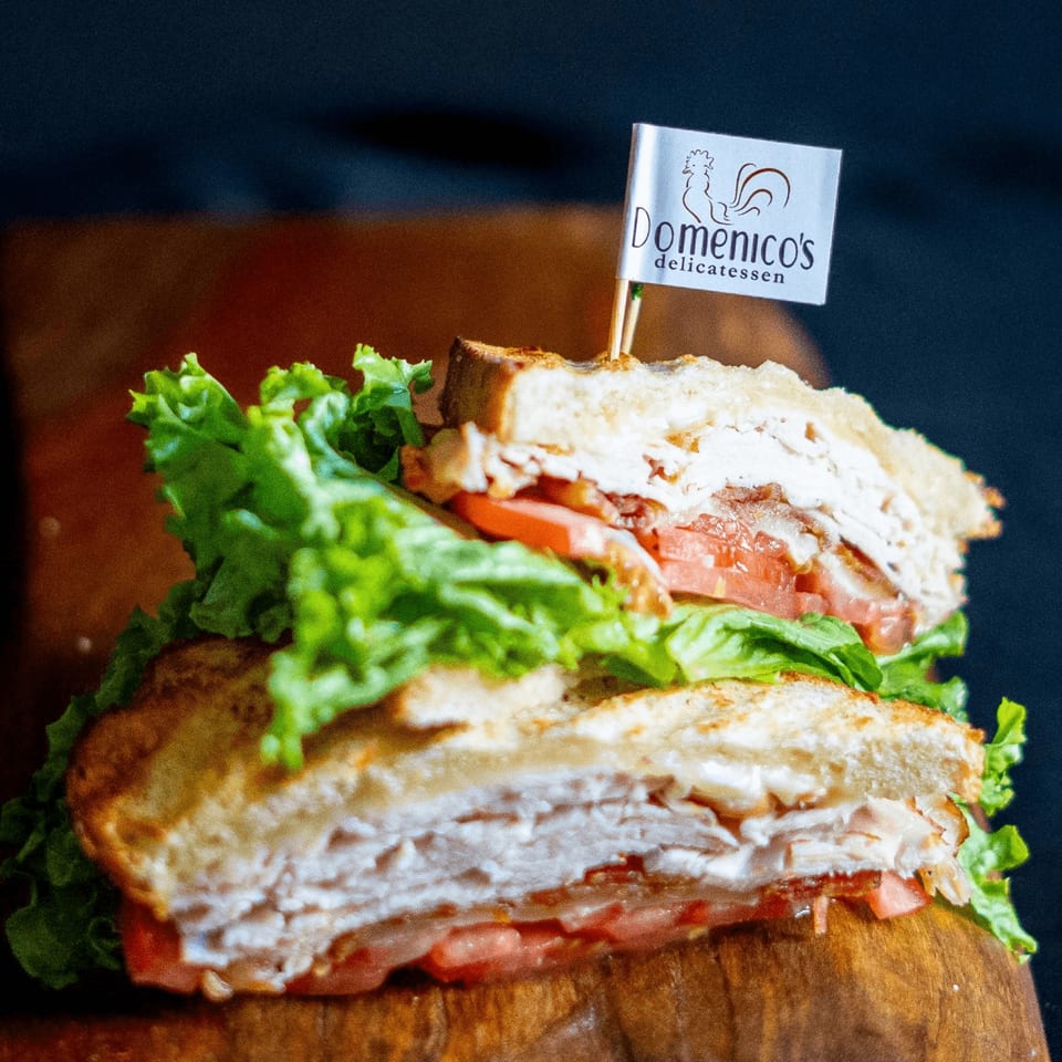Stacked deli sandwiches with lettuce, tomato, sliced meats, and creamy spread, topped with a Domenico’s Delicatessen flag.