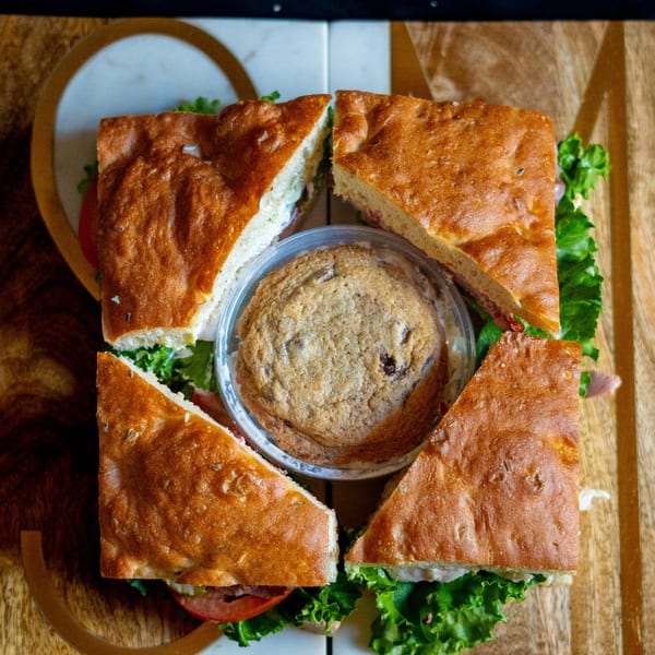 Four focaccia sandwich quarters surround a chocolate chip cookie on a wooden and marble surface.