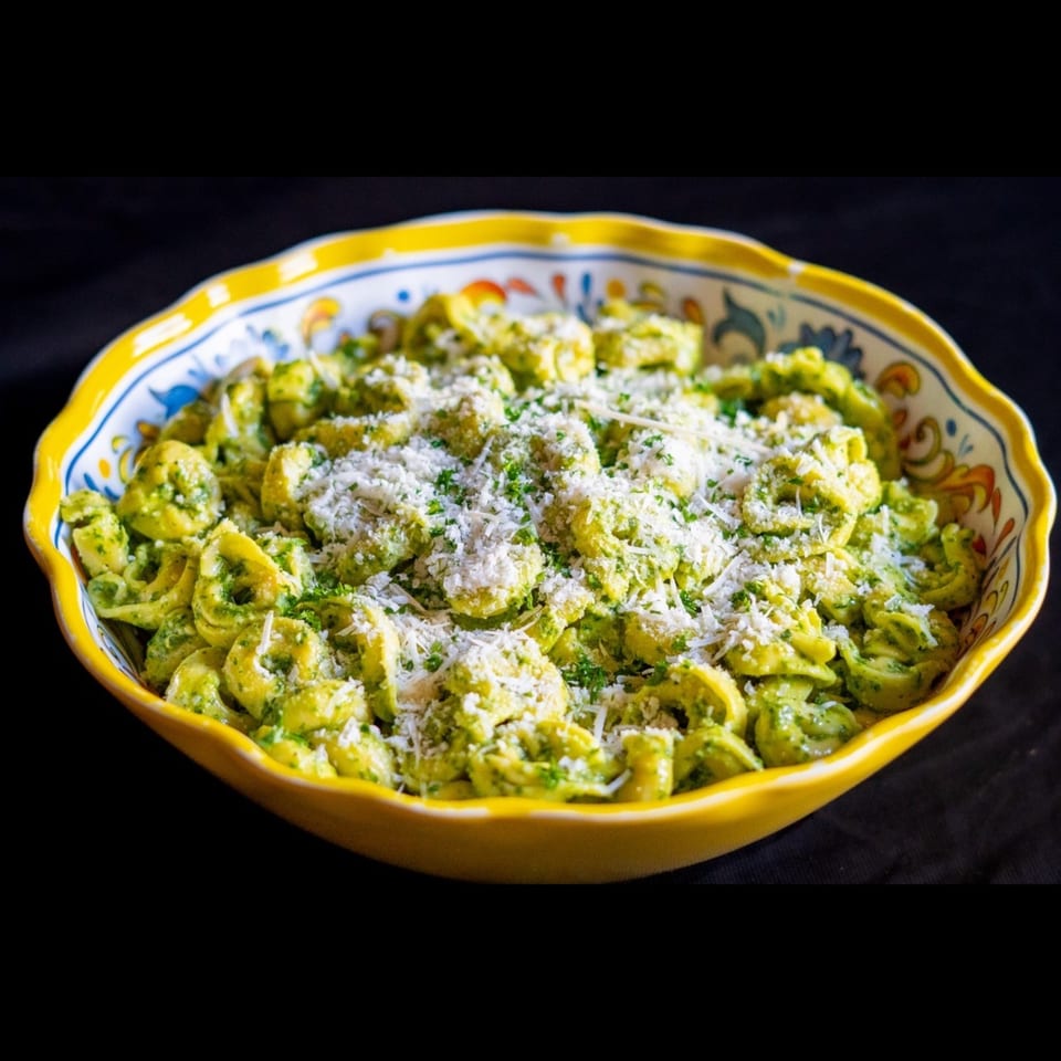 Tortellini pasta coated in green pesto, topped with grated cheese and herbs, served in a decorative yellow bowl.