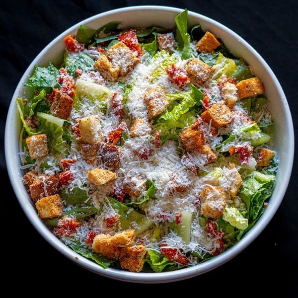 A close-up of a Caesar salad from Domenico's Delicatessen, topped with croutons and grated cheese.