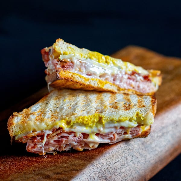 Two halves of a grilled Domenico's Delicatessen sandwich with melted cheese, ham, and mustard on a wooden board.