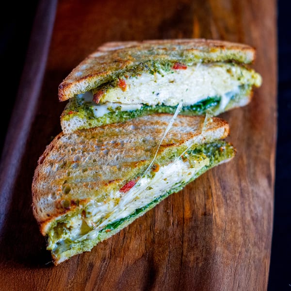 A grilled Domenico's Delicatessen sandwich, cut in half, with melted cheese and pesto on a wooden board.